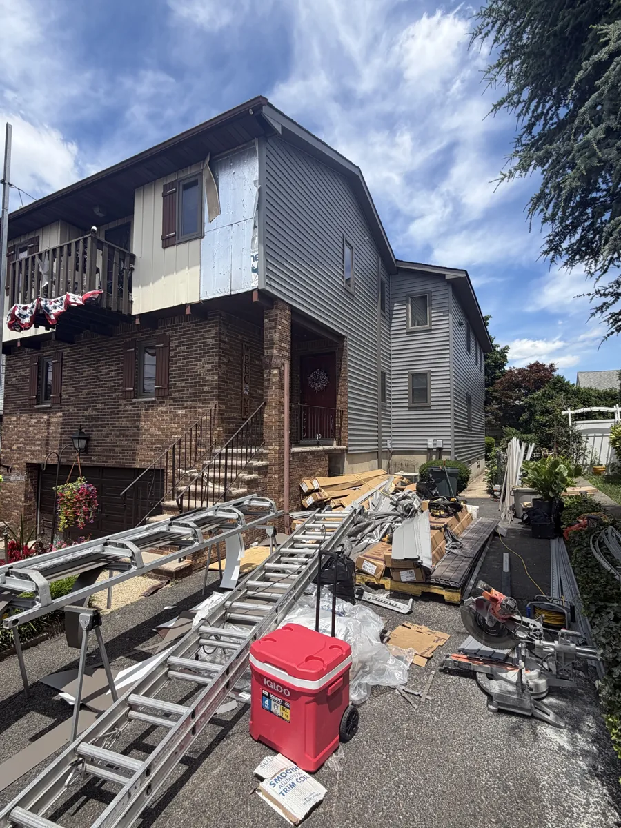 Two-story brick and frame house during siding replacement, with pump-jack staging and materials in the driveway