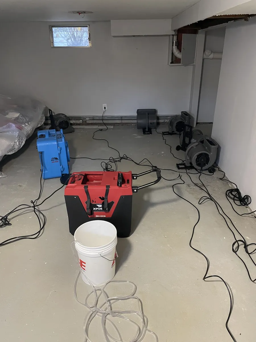 Basement with baseboards removed, a dehumidifier draining into a bucket, an air scrubber and air movers along the walls